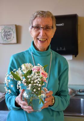 Short haired women with glasses in a blue sweater, smiling, and holding a vase of pink and white flowers.