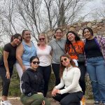 Eight women smile for a group photo on a casual hike on a cloudy day with six women standing in the back and two women squatting in the front wearing sunglasses.