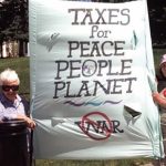 Six women outdoors on a sunny day wearing sunglasses and hats while standing next to and holding a large poster stating: Taxes for Peace People Planet.