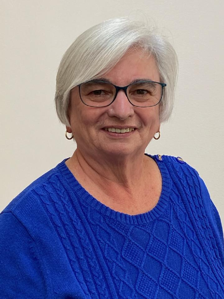 An older woman with a short grey bob smiles for a more formal photo. She's wearing a bright blue textured sweater, small hoop earrings an glasses.