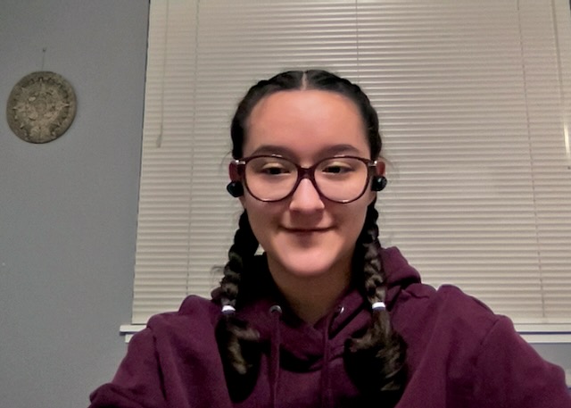 A student with big glasses and her hair in two braids, ear buds and a maroon sweatshirt looks into the camera.