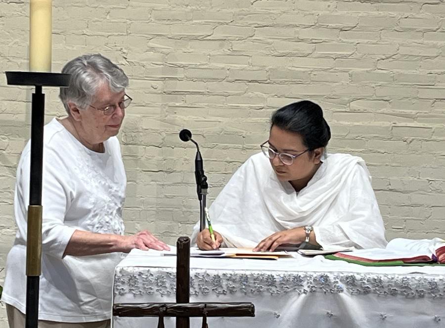 An older woman in white looks on as a younger woman with dark hair and traditional Pakistani clothing signs a document on an altar.