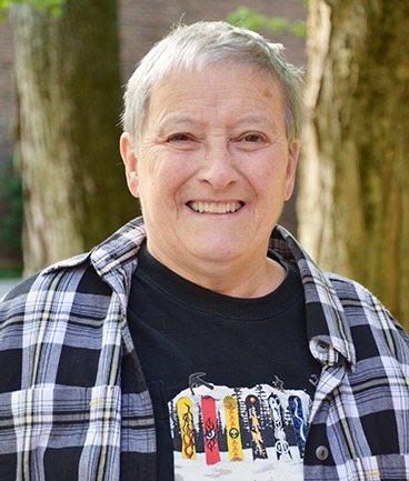 Photo of an older woman with short grey hair smiling, she's wearing a black t-shirt with a colorful patterned design on the front and a black and white flannel over it.