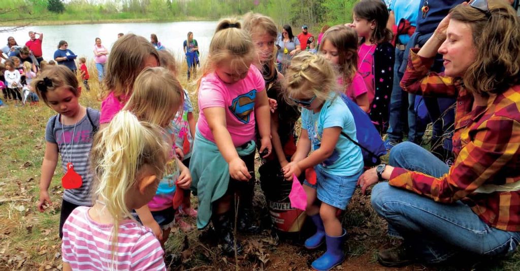 An adult woman squats down to be eye level with small children as they look inquisitively at a plant.