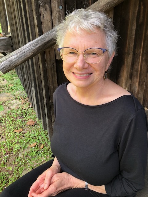 A white older woman with short grey hair smiles for the camera on a stoop outside a cabin. She's wearing large blue cat eye glasses, dangly earrings and a black outfit.