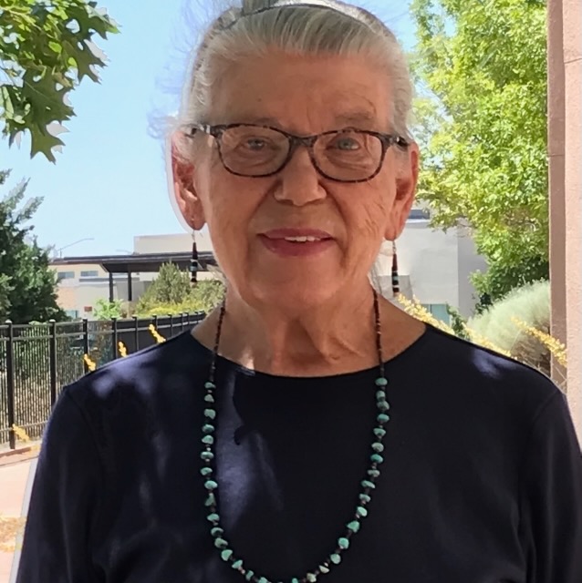 An older woman smiling on a sunny day. She is wearing a silver headband with her grey hair pulled back, tortoise shell glasses, dangly earrings, a blue shirt and long blue beaded necklace.