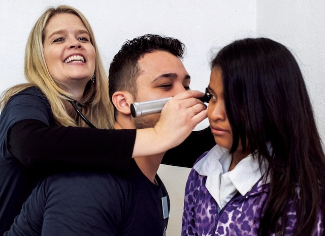 A pediatrician reaches over a student to show him how to examine an eardrum of a patient.