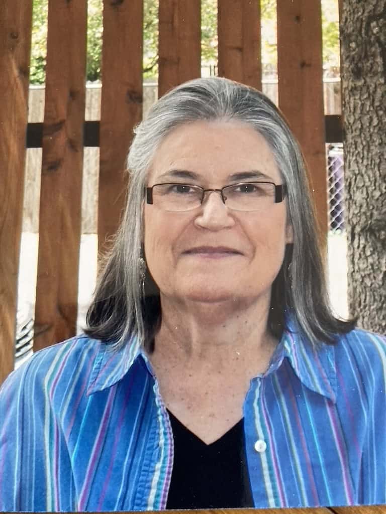 An older woman with shoulder length brown and grey hair smiles as she wears rectangular framed glasses, a black shirt underneath a blue striped collared shirt.