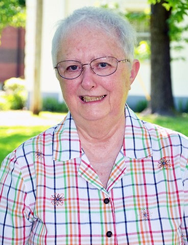 An older woman smiles for a camera on a sunny day outside. She is wearing rounded glasses and a bright pastel checkered shirt with embroidered flower details on it.