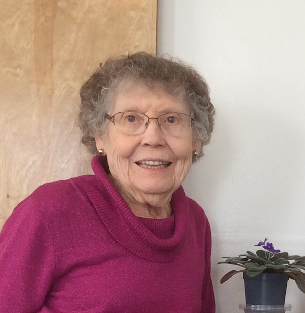 An older Sister of Loretto is seen smiling in front of a neutral backdrop with a plant next to her. She has grey an brown curly hair, silver earrings, rectangular glasses and a bright pink turtleneck.