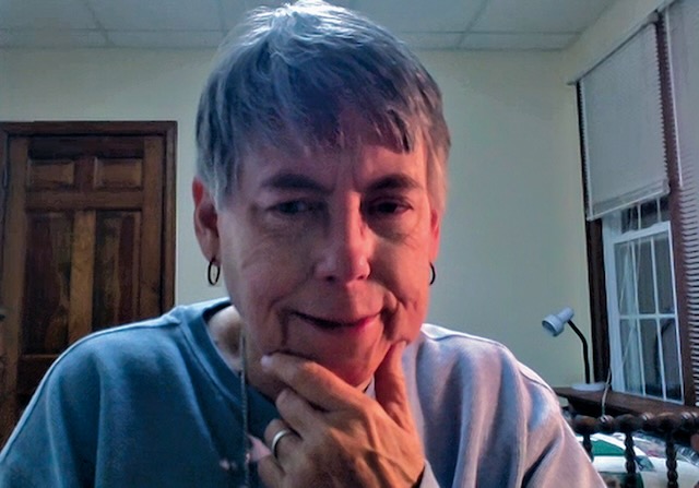 An older woman with short grey hair and small hoop earrings has her hand on her chin and looks inquisitively at the camera.