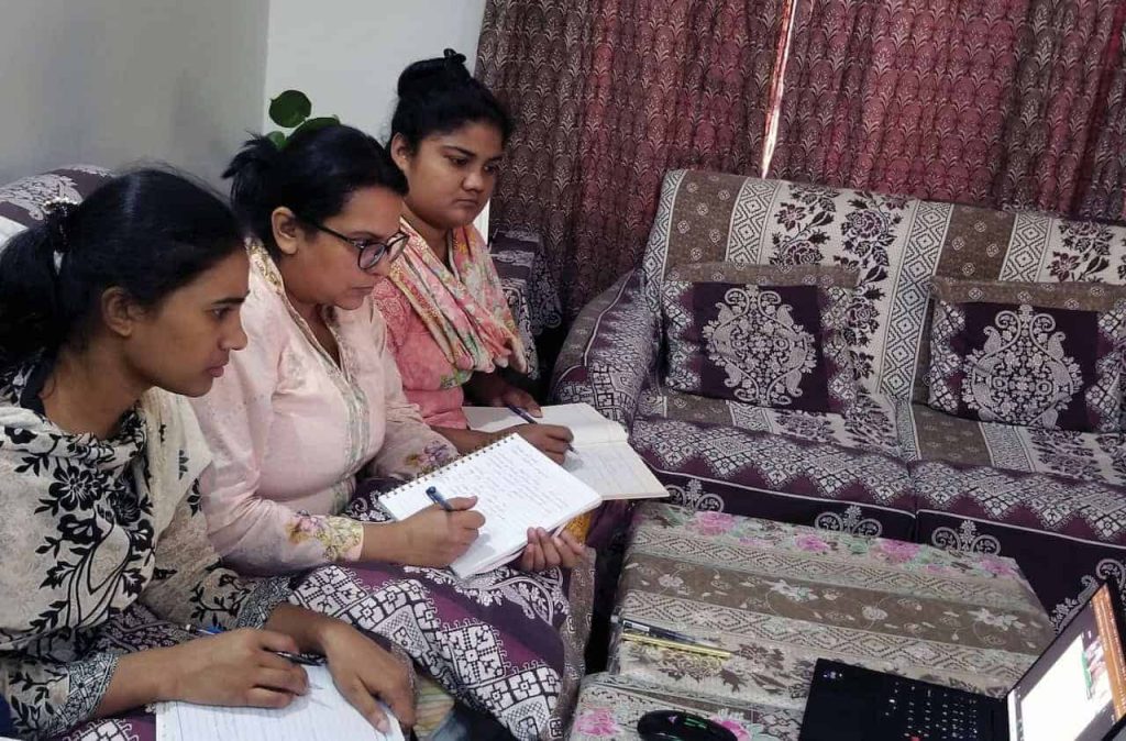 Three Pakistani women sitting on a colorful patterned purple couch with notebooks in their hands looking at a computer screen and studying intensely.