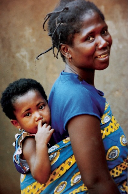 Photo of a Ghanaian mother and child. The child is held on the mother's back by a bright yellow and blue patterned wrap.