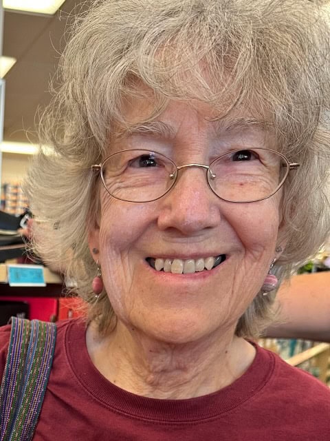 An older woman smiles close to the camera, she has grey wavy hair that frames her face, as well as metal round glasses, dangly earrings and a red t-shirt.