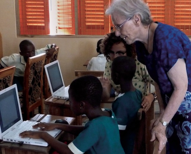 An older woman in a blue dress and glasses stands over a young student working on a computer to offer support.