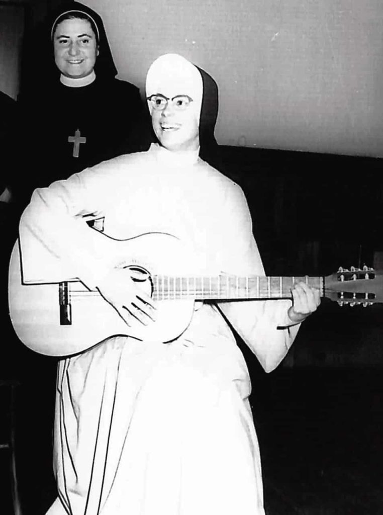 A black and white photo of a nun in habit smiling and strumming on guitar. Another sister in habit stands behind her enjoying the performance.