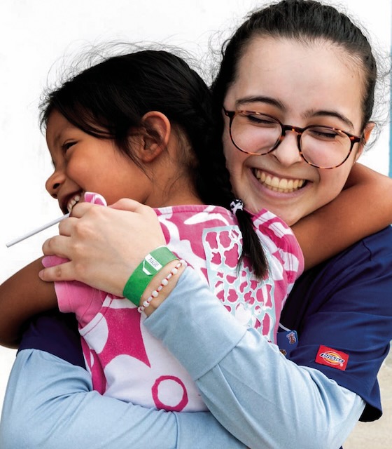 A pre-med student in blue scrubs smiles while sharing a hug with a small child in a pink flowered t-shirt.