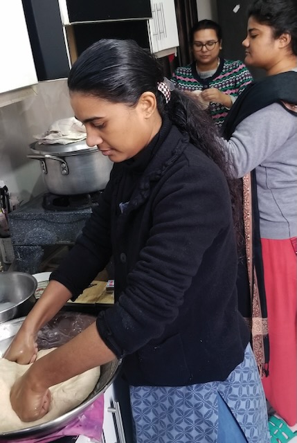 Three pakistani women are shown making break in a kitchen. The woman closest to the camera is shown kneading the bread with the other two form the break in the background.