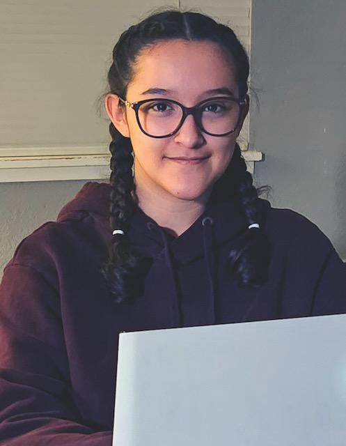 A high school student with big black round glasses and braids smiles softly while wearing a dark purple sweatshirt