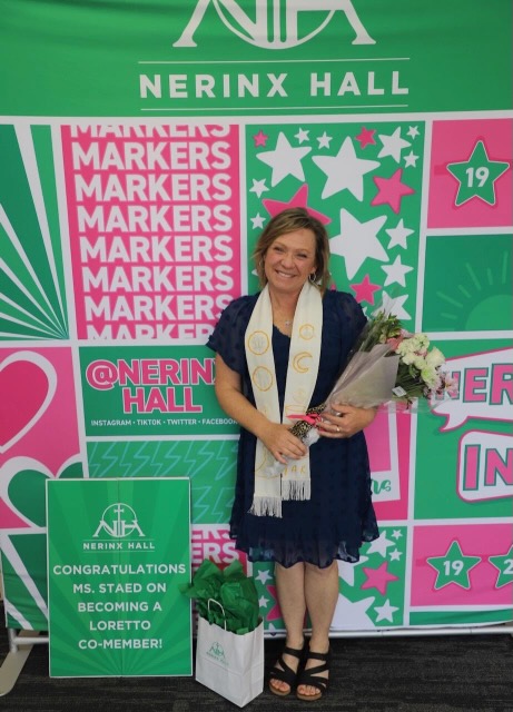 A woman is smiling and holding a bouquet of flowers standing in front of a bright green and pink banner that says Nerinx Hall on it.