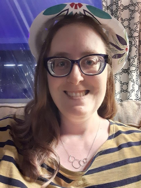 Photo of a woman smiling with thick glasses, a colorful hat, mid length light brown hair wearing a striped black and yellow t-shirt and a silver necklace with multiple cirlces on it.