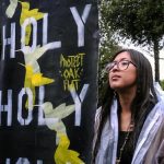 A young indigenous girl with big glasses, long dark hair and a necklace with a shell on it stands powerfully holding a sign that says "Holy Holy Holy Holy Protect Oak Flat."