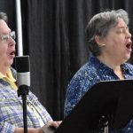 Two women sing their hearts out in front of microphones and music stands.