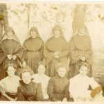 Archival photo of school personnel - four habited nuns pose in the back row, with five younger women in dresses seated in a row in front.