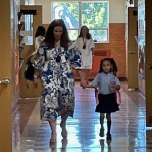 A school staff member walks down the hallway hand in hand with a child wearing a backpack.