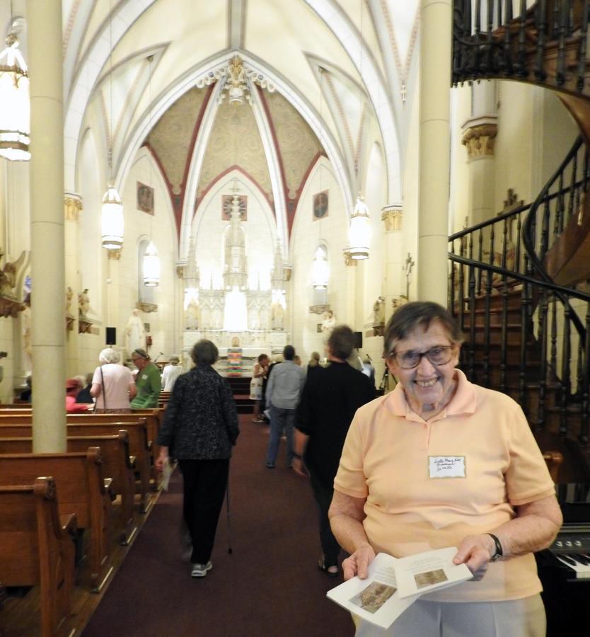 A woman hands out programs and warm smiles as you enter a beautiful church. A winding staircase is visible behind her to the right.