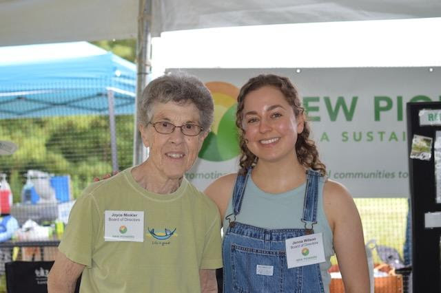 From left, an older woman with grey hair, glasses and a green t-shirt stands smiling with a young woman with brown curly hair wearing a blue tank top and jean overalls. They are standing together in front of a sign that says "new pioneers for a sustainable future."