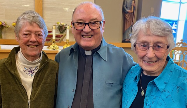 Three older Loretto members smile together for a group photo. On the left there is a woman with grey hair, a white turtleneck and green jacket, in the middle a priest, with black framed glasses and a light blue jacket and on the right is a woman with grey short hair, clear framed rectangle glasses wearing a black shirt, gold necklace and blue flower patterned shirt over the black shirt.