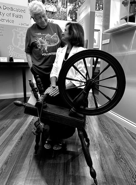 A black and white but current photo of an older woman looking down at another woman in a white blazer as she sits on a spinning wheel.