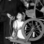 A black and white photo of a nun in habit looks down at a small child in a white dress as she shows her a spinning wheel.