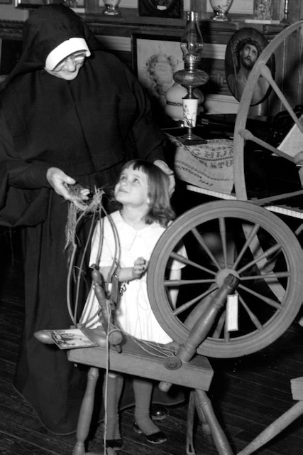 A black and white photo of a nun in habit looks down at a small child in a white dress as she shows her a spinning wheel.