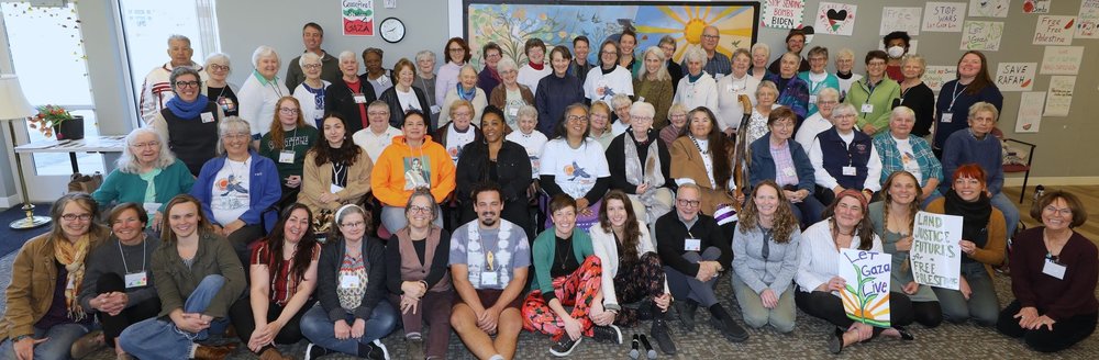 A large group photo with multiple rows of people standing and sitting, all smiling together indoors. Two sitting are holding posters similar to those on the wall behind them