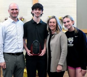 A tall male teenager with black floppy hair and a black t-shirt and jeans stands with his father (left), mother and sister (right) smiling as he holds a glass award.