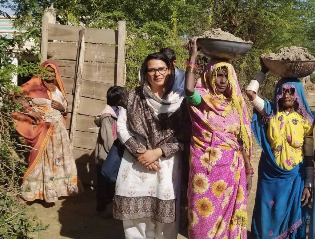 A group of Pakistani women outside wearing colorful patterned clothing. A woman in glasses wearing a patterned brown and white outfit looks straight at the camera. She is surrounded by women who are carrying mud in bowls on their heads.