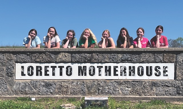 Eight students in a row leaning over a sturdy rock sign that says "Loretto Motherhouse" in black text and a white background