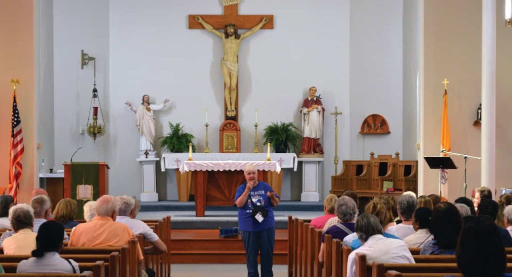 An older white woman with white hair and a blue shirt holds a small microphone and is shown speaking to a crowd in the pews in front of the altar of a Catholic Church.