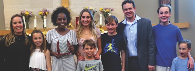 A family photo with two parents and seven children of varying ages smiling as their teenage daughter holds an award she just received.