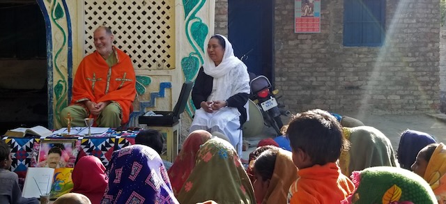 An Irish priest wearing orange and green attire sitting next to a nun wearing a black outfit and a white shawl covering her head, speaking to a group of people during a Mass.