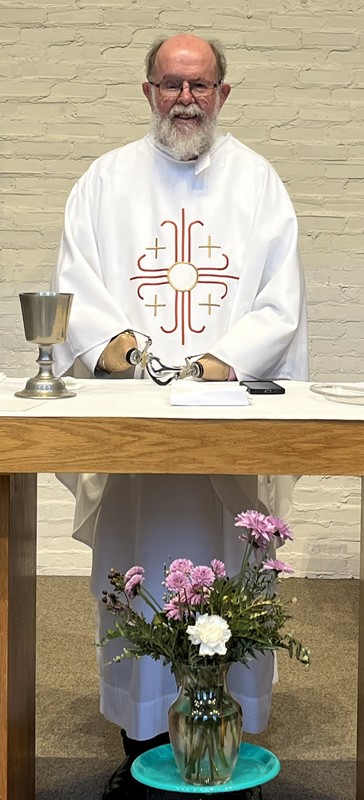 A priest smiling for a picture while standing in front of a table on stage at church.