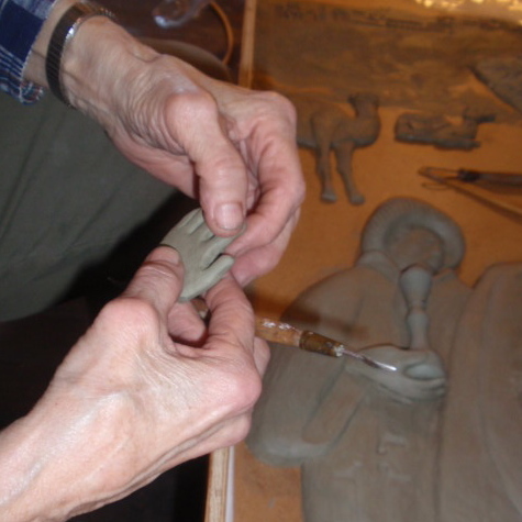 A pair of hands shape a clay hand to go on a sculpture, visible laying on a workspace in the background