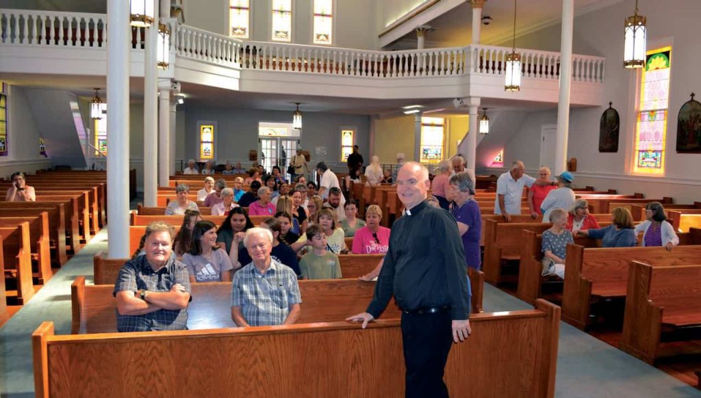 A white male pastor in black clothing smiles as he speaks with guests in the pews of his church.