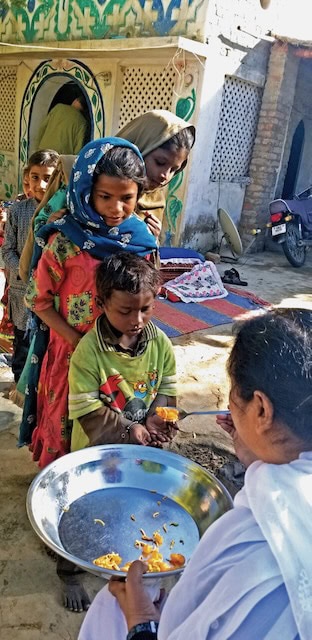 Children in colorful clothing line up outside to receive sweets from a religious sister dressed in white.