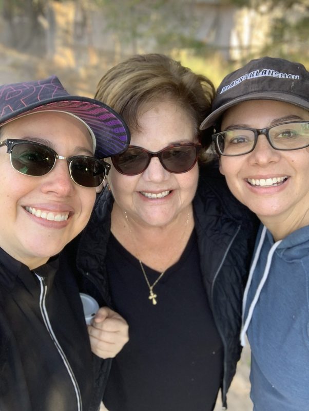 Three women smile together closely for a group selfie outdoors on a sunny day.