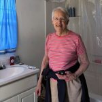 An older white woman with grey hair and round framed glasses, a striped pink shirt and beige pants smiles in a newly constructed and completed bathroom.