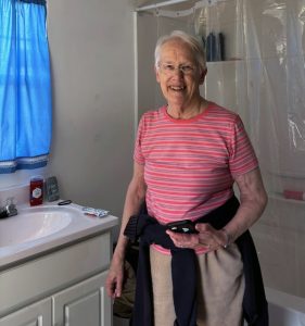 An older white woman with grey hair and round framed glasses, a striped pink shirt and beige pants smiles in a newly constructed and completed bathroom.