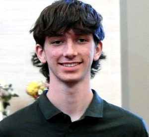 A tall male teenager with black floppy hair and a black t-shirt and jeans stands smiling as he holds a glass award.